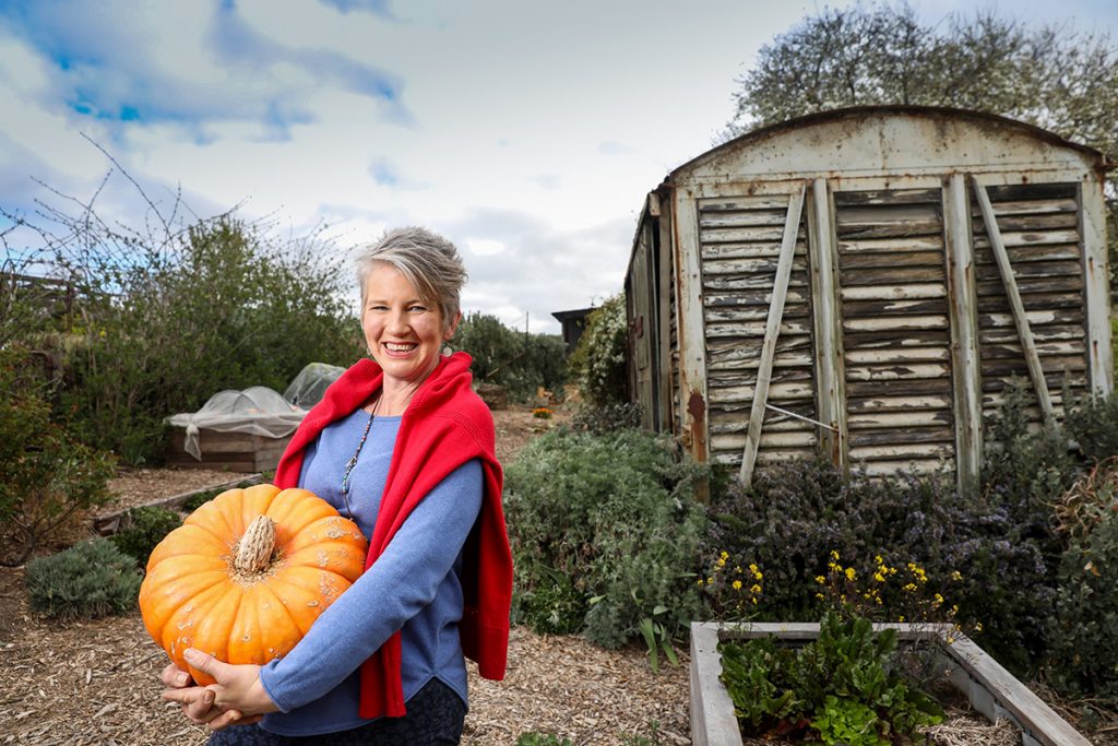 Smiling woman holding large pumpkin in front of sustainable garden